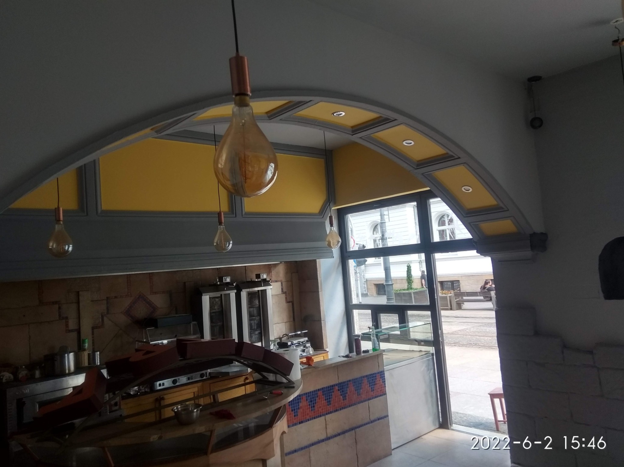 Wnętrze lokalu gastronomicznego z widoczną kuchnią, wykończeniem ścian w mozaikę i dekoracyjnym łukiem z żółtymi panelami pod sufitem, oświetlone wiszącymi żarówkami w stylu retro.