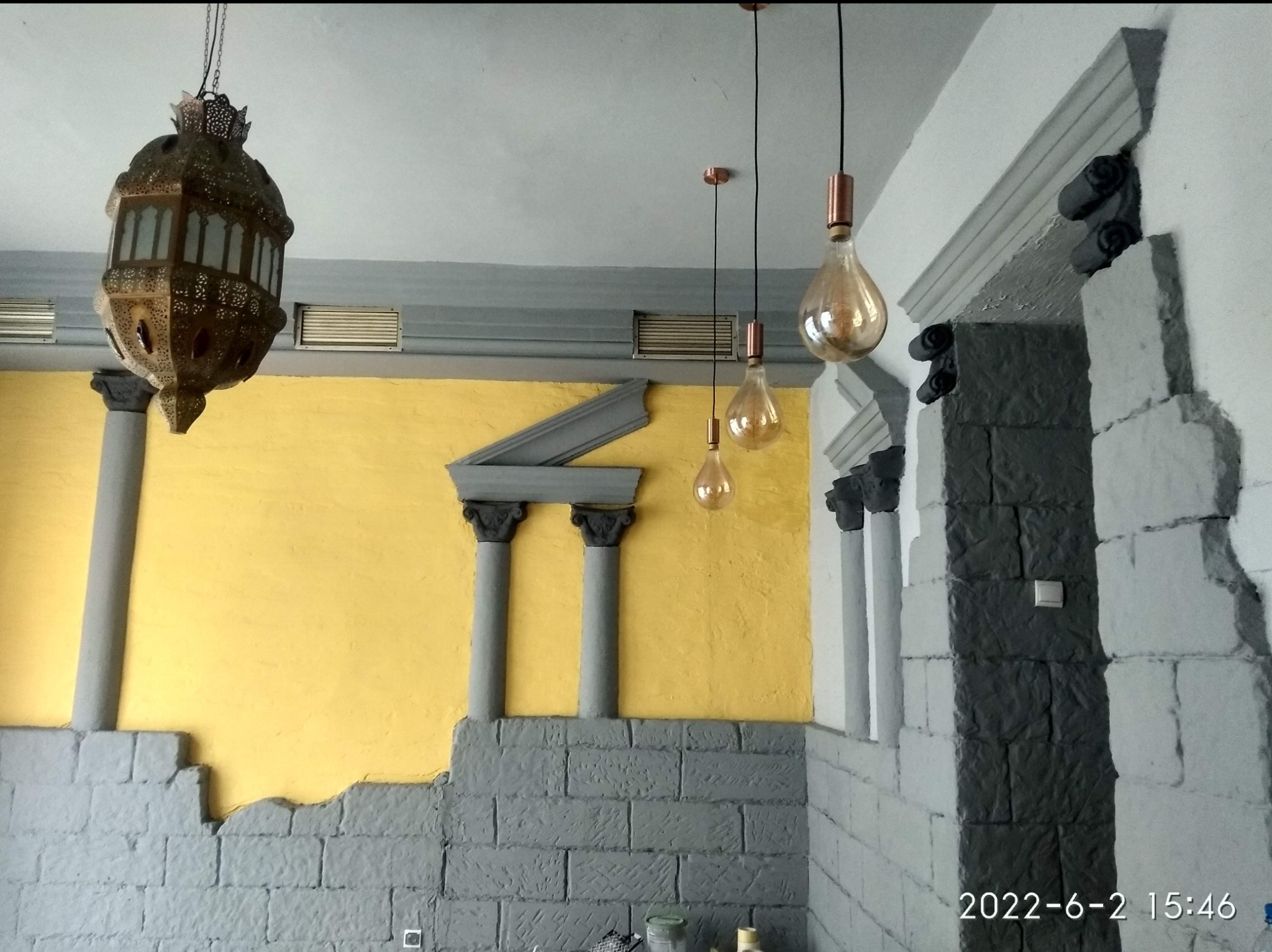 Żółta ściana z imitacją kolumn i cegieł, szara dekoracyjna listwa pod sufitem, ozdobna lampa wisząca w stylu marokańskim oraz trzy żarówki dekoracyjne na tle białej ściany z imitacją kamiennej ściany.