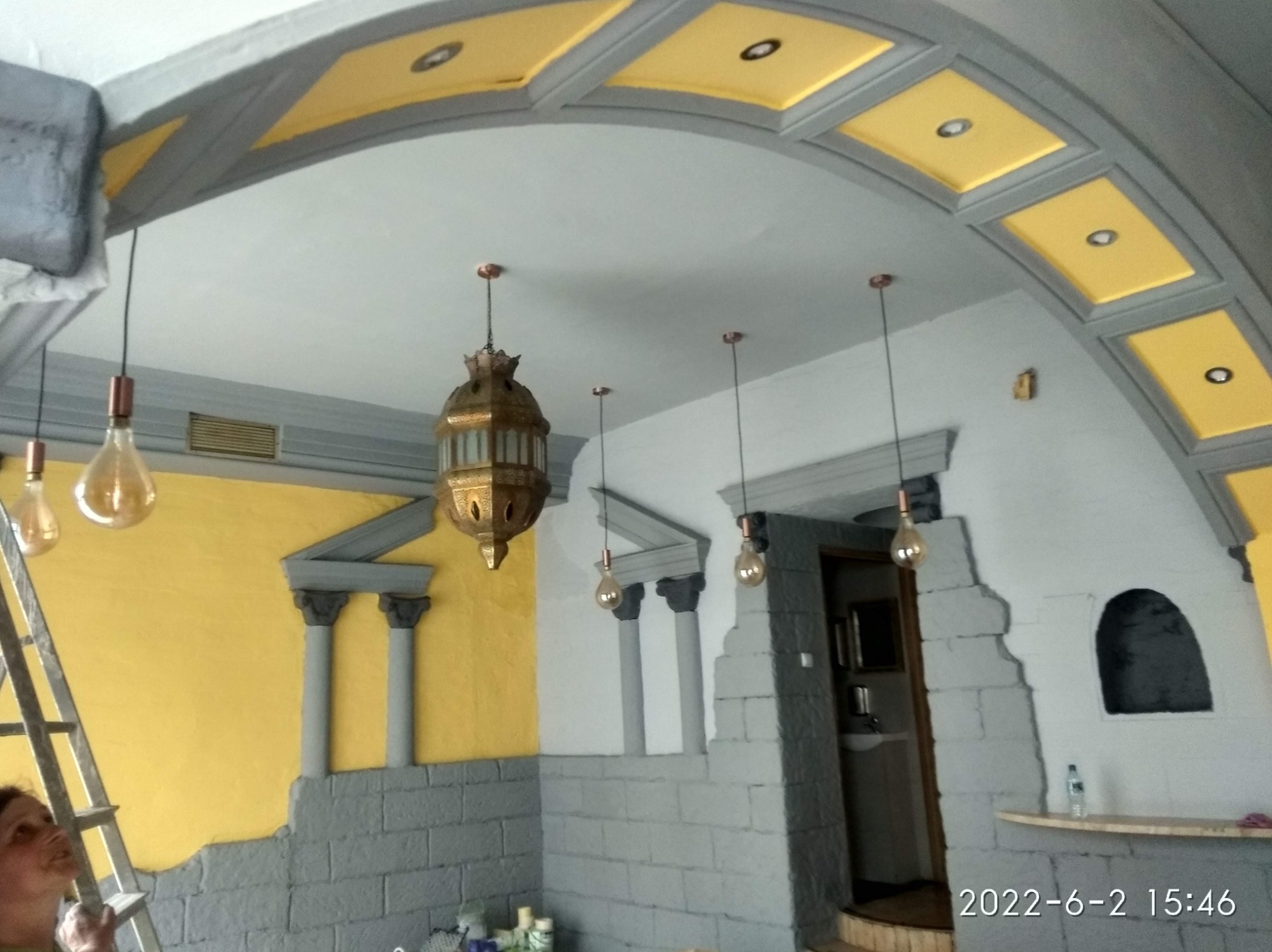 Stylizowane wnętrze z żółto-szarą kolorystyką, imitacją kolumn i kamiennej ściany, ozdobnymi lampami wiszącymi i dekoracyjnym sufitem z żółtymi panelami