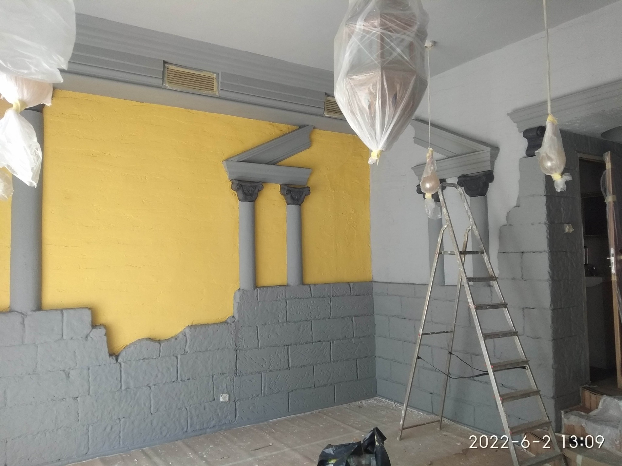 Ściana z dekoracyjnym malowaniem imitującym starożytne kolumny i mur, częściowo w kolorze żółtym, częściowo szarym, z zabezpieczonymi lampami wiszącymi i drabiną w pomieszczeniu w trakcie prac...
