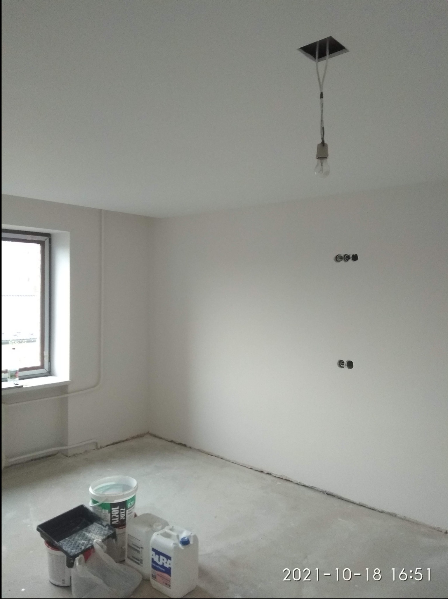 Pokój w trakcie remontu: świeżo pomalowane białe ściany, widoczne puszki z farbą i narzędzia malarskie na podłodze, surowa żarówka zwisająca z sufitu, gniazdka elektryczne bez osprzętu.