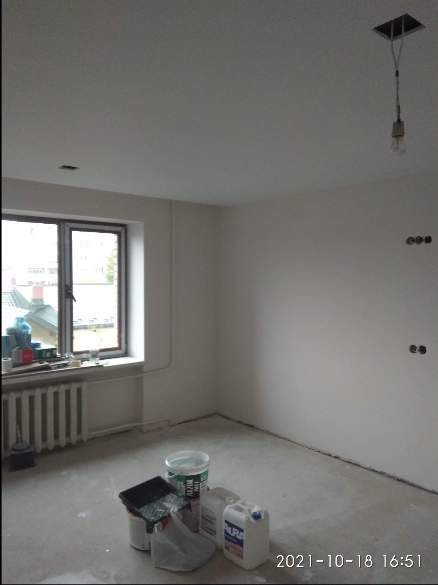 Pomalowane na biało wnętrze pokoju z widocznymi narzędziami malarskimi, grzejnikiem i oknem z widokiem na zewnątrz. Na suficie widoczny otwór po lampie z wiszącą żarówką.