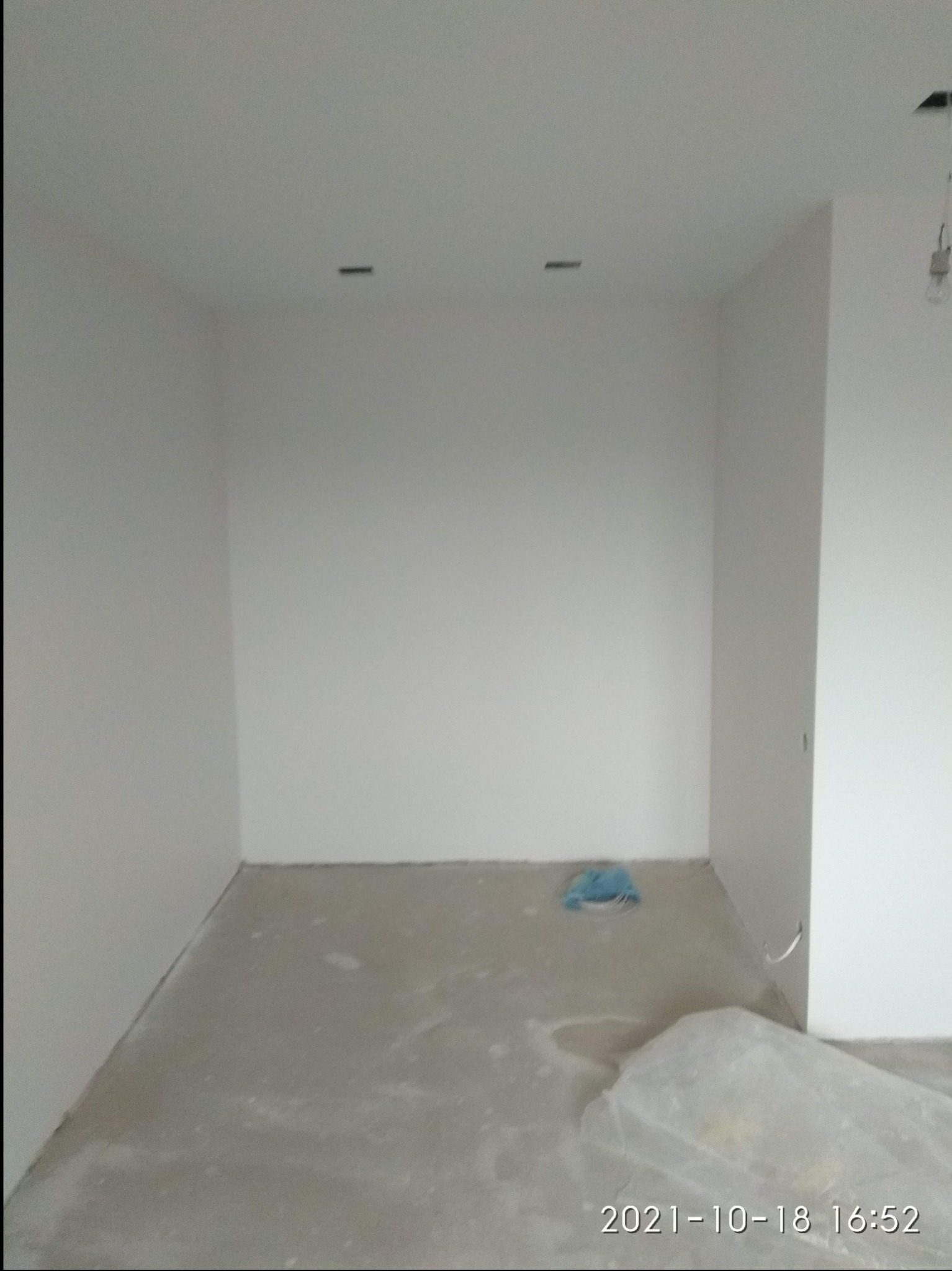 Pusty pokój po pracach przygotowawczych, ściany pomalowane na biało, surowa podłoga z widocznym zabezpieczeniem w rogu, brak lampy.