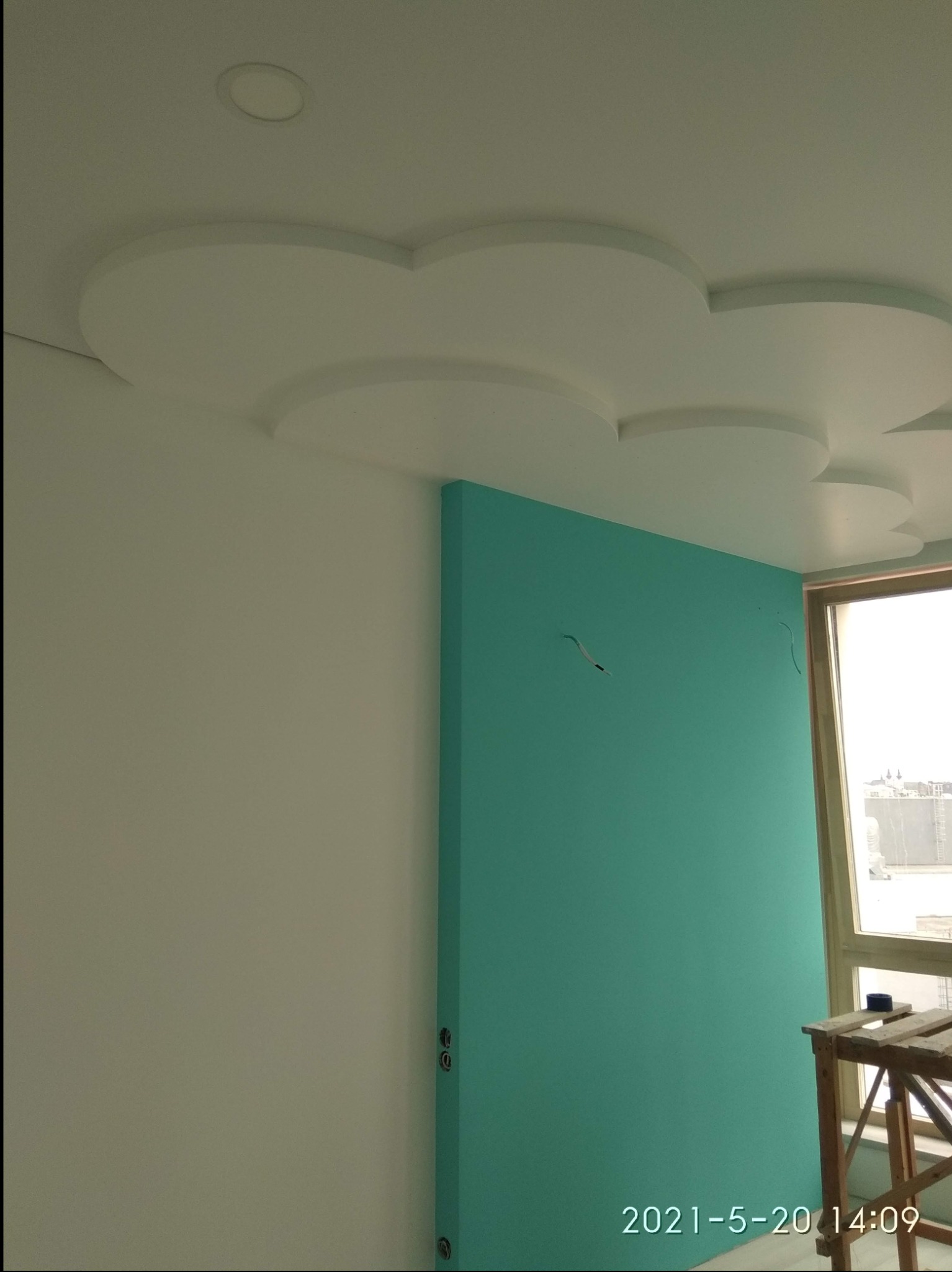 Ściana pomalowana na turkusowo w pokoju z dekoracyjnym sufitem w kształcie chmur, widoczne wystające przewody i drabina przy oknie.