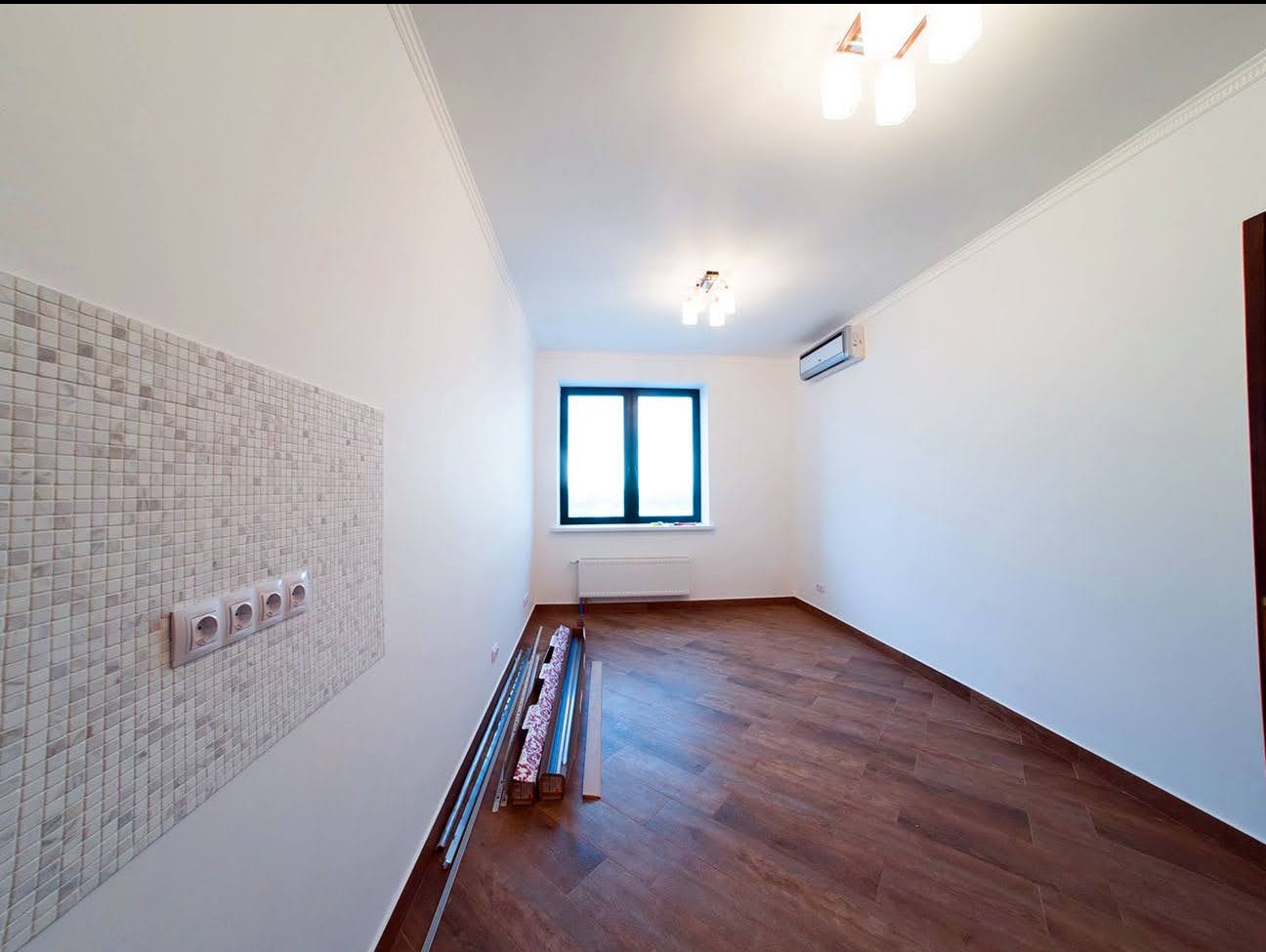 Wnętrze pokoju w Chorzowie po remoncie: gładkie, białe ściany z listwami dekoracyjnymi pod sufitem, brązowa podłoga, okno z czarną ramą oraz fragment ściany wyłożony mozaiką z gniazdkami...