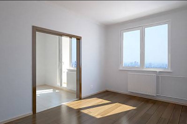 Jasne wnętrze pokoju z drewnianą podłogą, białymi ścianami i grzejnikami pod oknami, widoczne przejście do kolejnego pomieszczenia.