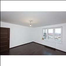 Pusty pokój po remoncie: gładkie, białe ściany, ciemna podłoga, brązowe drzwi, okno z widokiem na miasto, centralnie umieszczona lampa sufitowa.