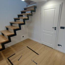 MasterArt - Nowoczesne schody o konstrukcji stalowej z drewnianymi stopniami, zamontowane przy białej ścianie i drzwiach w jasnym wnętrzu. Na podłodze widoczne narzędzia i elementy montażowe.