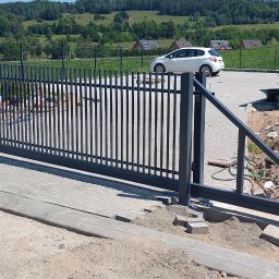 MasterArt - Nowoczesna, przesuwna brama wjazdowa z metalowymi sztachetami, widoczna w trakcie montażu na tle zielonego krajobrazu. Częściowo ułożona kostka brukowa.