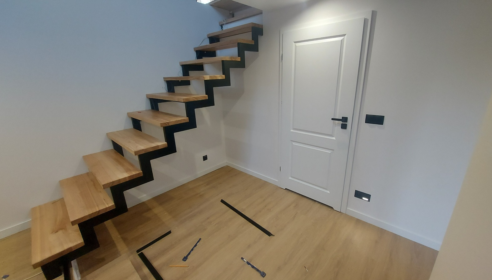 Nowoczesne schody o konstrukcji stalowej z drewnianymi stopniami, zamontowane przy białej ścianie i drzwiach w jasnym wnętrzu. Na podłodze widoczne narzędzia i elementy montażowe.