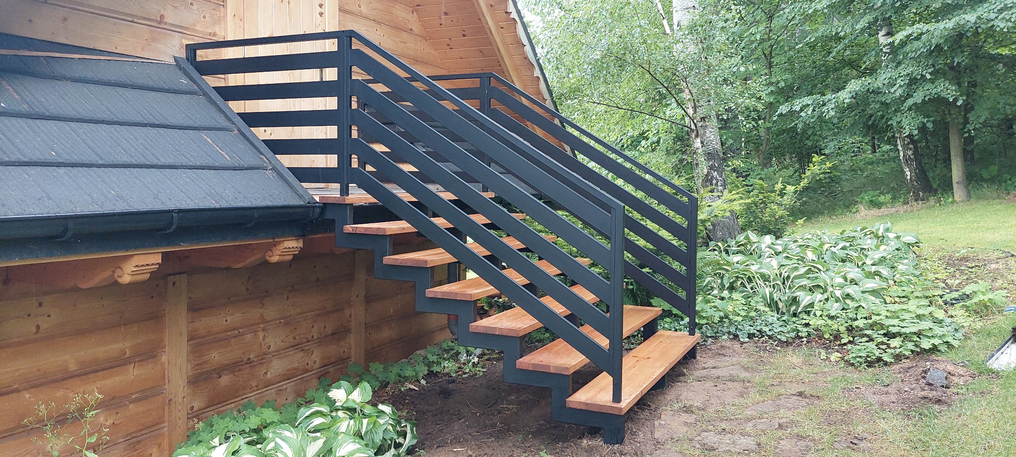 Zewnętrzne schody z metalową balustradą, drewniane stopnie, na tle drewnianego domu i zieleni. Ujęcie z dołu, podkreślające solidną konstrukcję i estetyczne wykończenie.