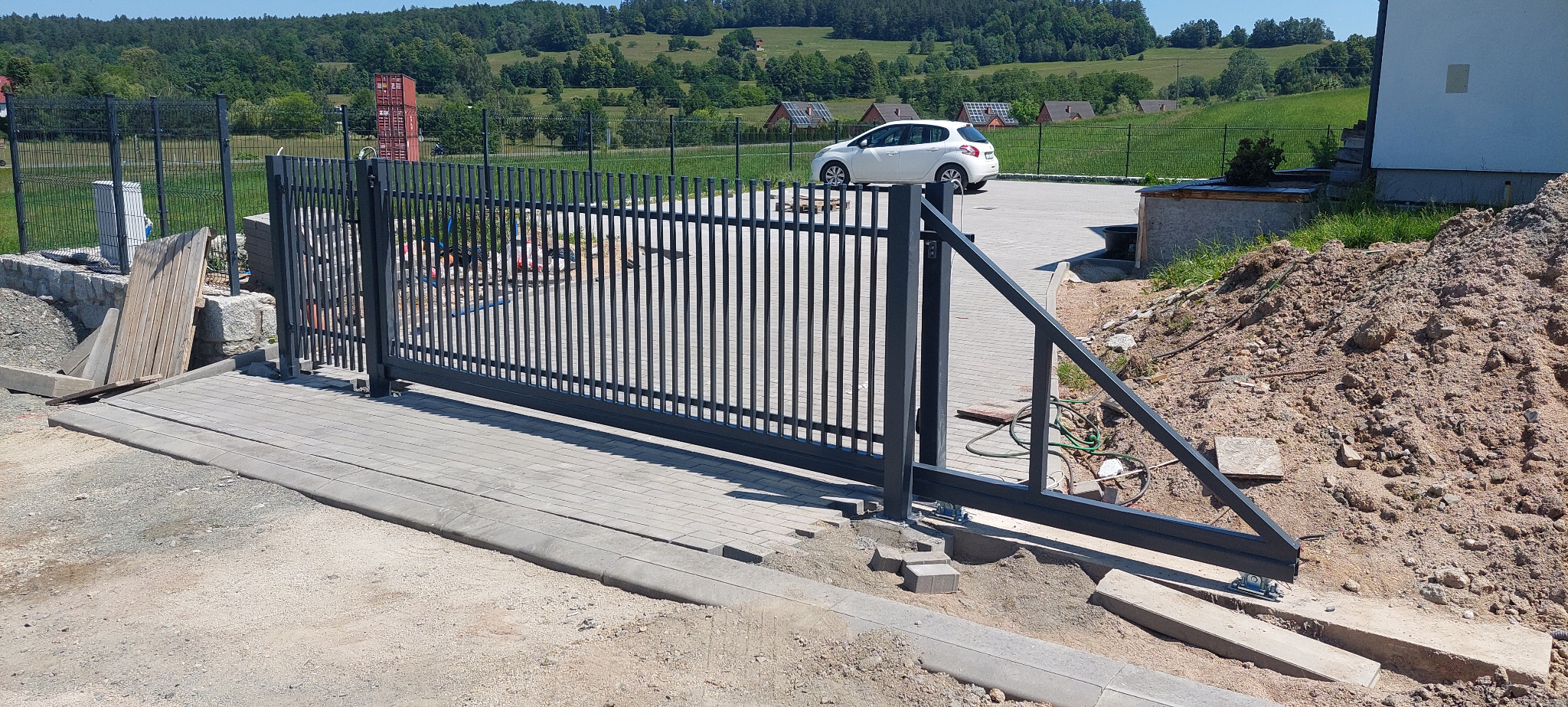 Nowoczesna, przesuwna brama wjazdowa z metalowymi sztachetami, widoczna w trakcie montażu na tle zielonego krajobrazu. Częściowo ułożona kostka brukowa.