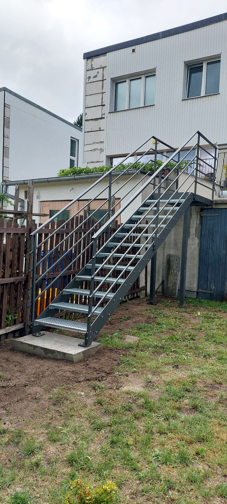 Zewnętrzne schody metalowe z kratowymi stopniami prowadzące do budynku. Szara konstrukcja na betonowej podstawie, widok z dołu, trawa i budynki w tle.