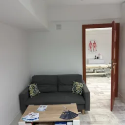 Poczekalnia gabinetu fizjoterapii z szarą sofą, stolikiem kawowym z ulotkami i otwartymi drzwiami do sali zabiegowej z leżanką i plakatem anatomicznym.