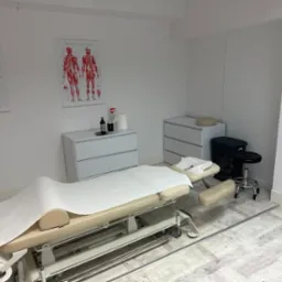 Jasne wnętrze gabinetu z regulowanym łóżkiem do zabiegów, pokrytym jednorazowym prześcieradłem, widoczne plakaty anatomiczne na ścianie, szafki i stołek na kółkach.