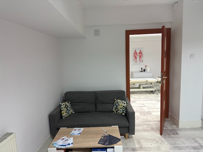 Poczekalnia gabinetu fizjoterapii z szarą sofą, stolikiem kawowym z ulotkami i otwartymi drzwiami do sali zabiegowej z leżanką i plakatem anatomicznym.