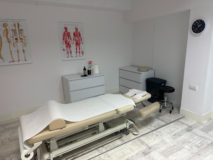 Jasne wnętrze gabinetu z regulowanym łóżkiem do zabiegów, pokrytym jednorazowym prześcieradłem, widoczne plakaty anatomiczne na ścianie, szafki i stołek na kółkach.