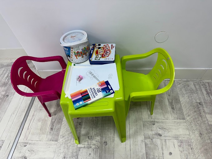 Dwa krzesła dziecięce (różowe i zielone) ustawione przy zielonym stoliku, na którym leżą kredki, kartka z narysowaną gitarą i temperówka, obok stoi wiaderko z zabawkami i gra planszowa.