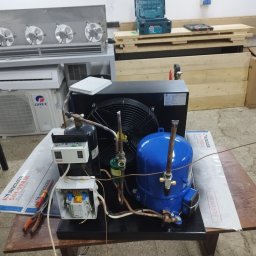ASKiD sp. z o.o. - Sprężarka chłodnicza z wentylatorem na stole warsztatowym, obok narzędzia i elementy instalacji. Widoczny klimatyzator Gree i opakowania Sinclair.