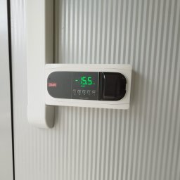 ASKiD sp. z o.o. - Panel sterowania Danfoss Optima Control z wyświetlaną temperaturą -15.5°C, zamontowany na ścianie chłodni z charakterystyczną, pionową fakturą.