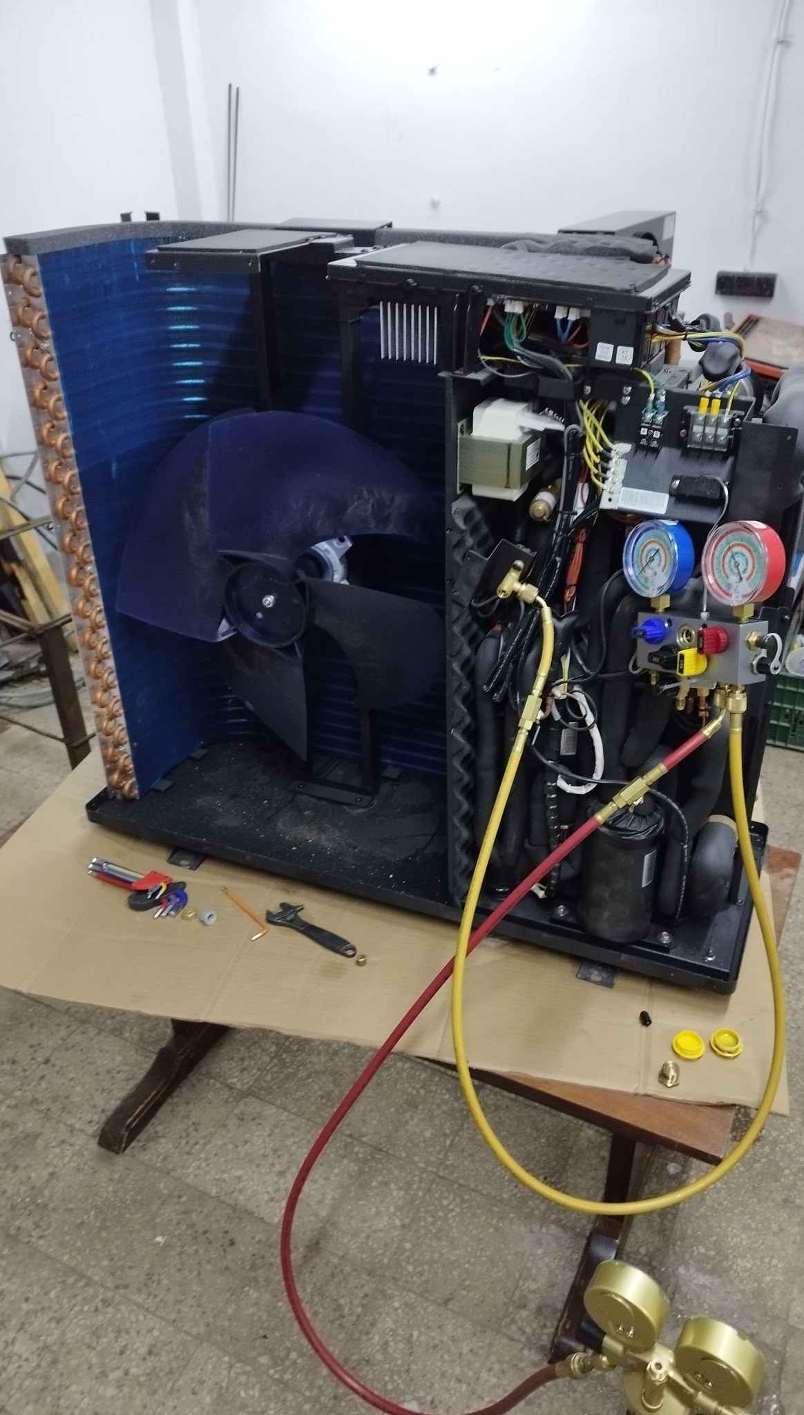 Otwarta obudowa klimatyzatora z widocznymi elementami wewnętrznymi i podłączonymi przewodami pomiarowymi. Na stole leżą narzędzia.