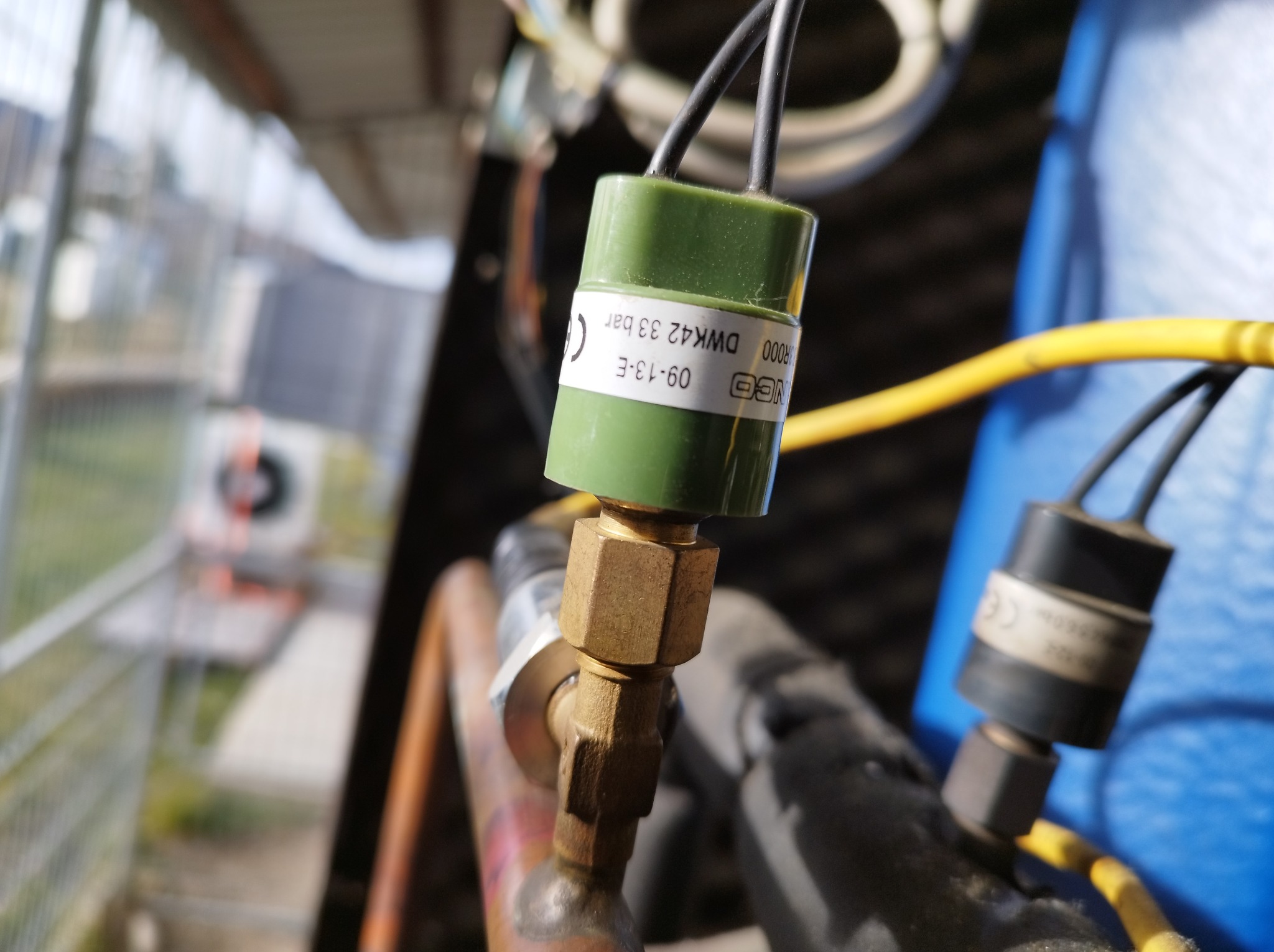 Szczegółowy widok na element instalacji chłodniczej z zielonym czujnikiem ciśnienia i mosiężnym zaworem, otoczone przewodami i elementami systemu.