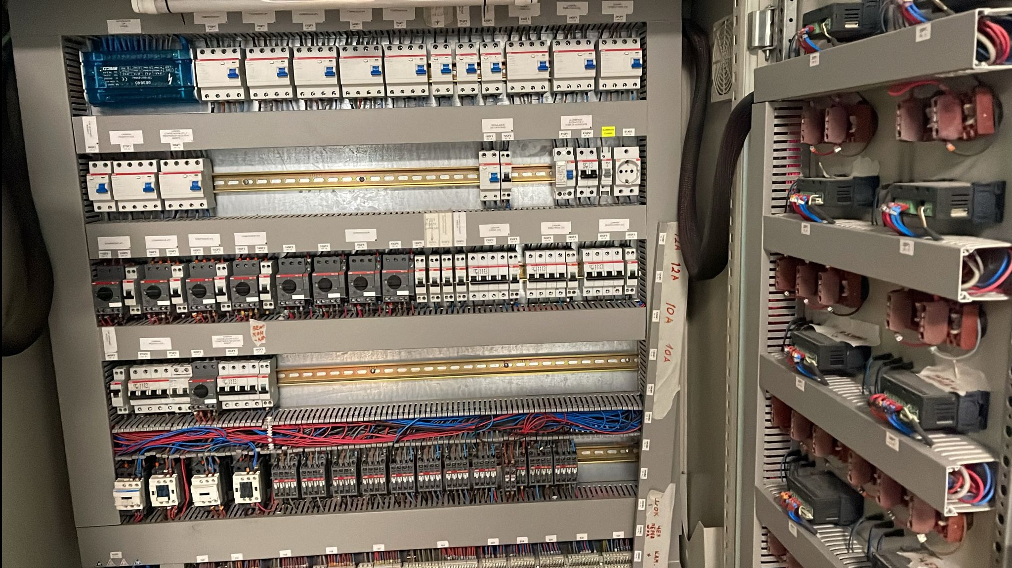 Szara szafa sterownicza z licznymi bezpiecznikami, przekaźnikami i okablowaniem, widoczne elementy instalacji elektrycznej i automatyki, rozmieszczenie komponentów.