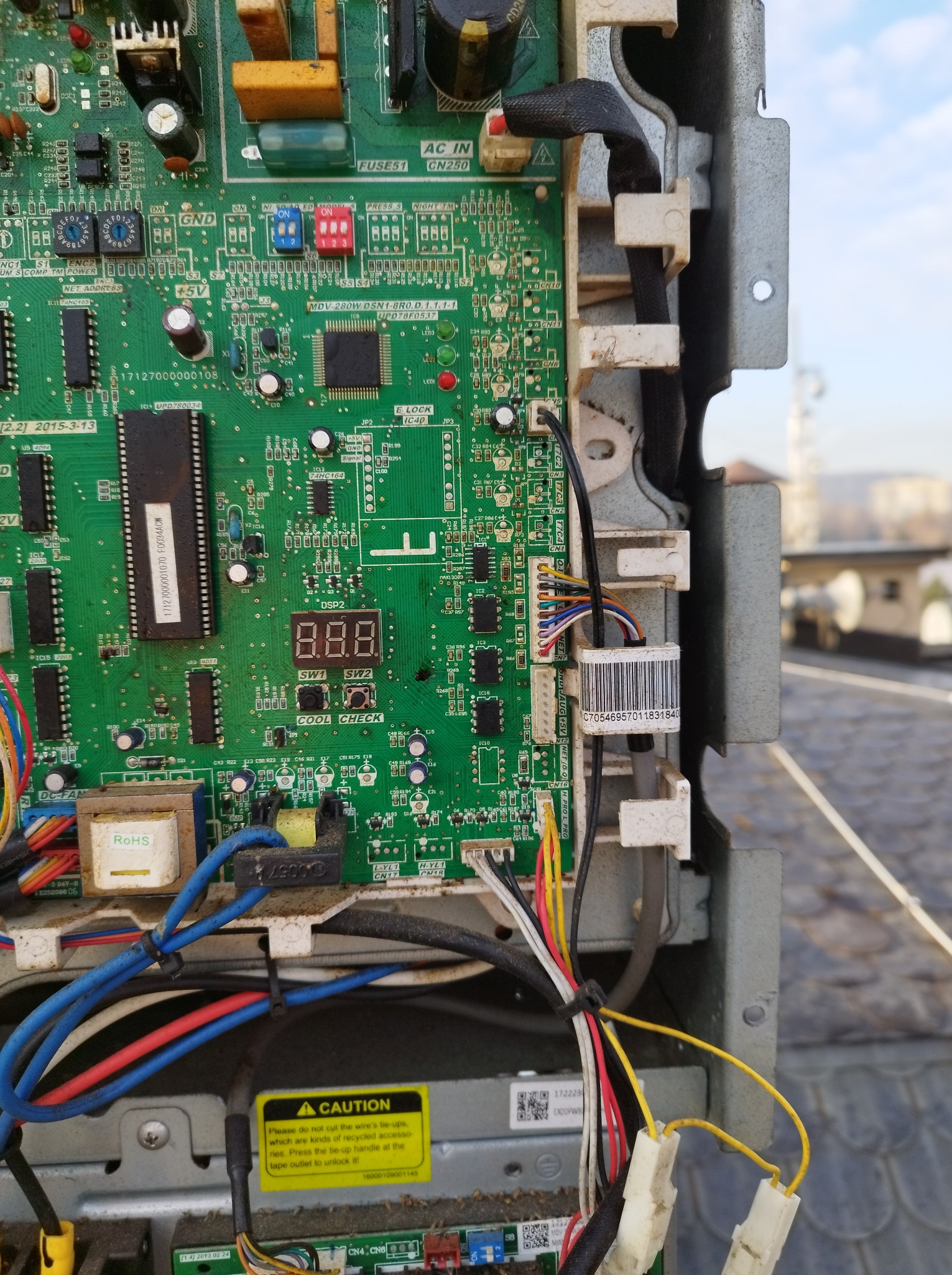 Wnętrze klimatyzatora z widoczną zieloną płytą główną, licznymi elementami elektronicznymi, przewodami i żółtą naklejką ostrzegawczą. Widoczne złącza i układy scalone.