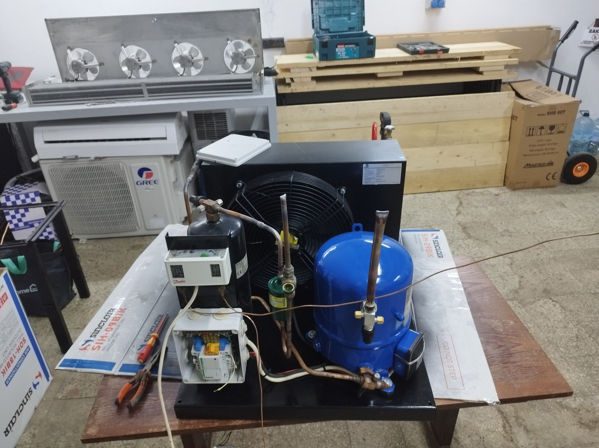 Sprężarka chłodnicza z wentylatorem na stole warsztatowym, obok narzędzia i elementy instalacji. Widoczny klimatyzator Gree i opakowania Sinclair.