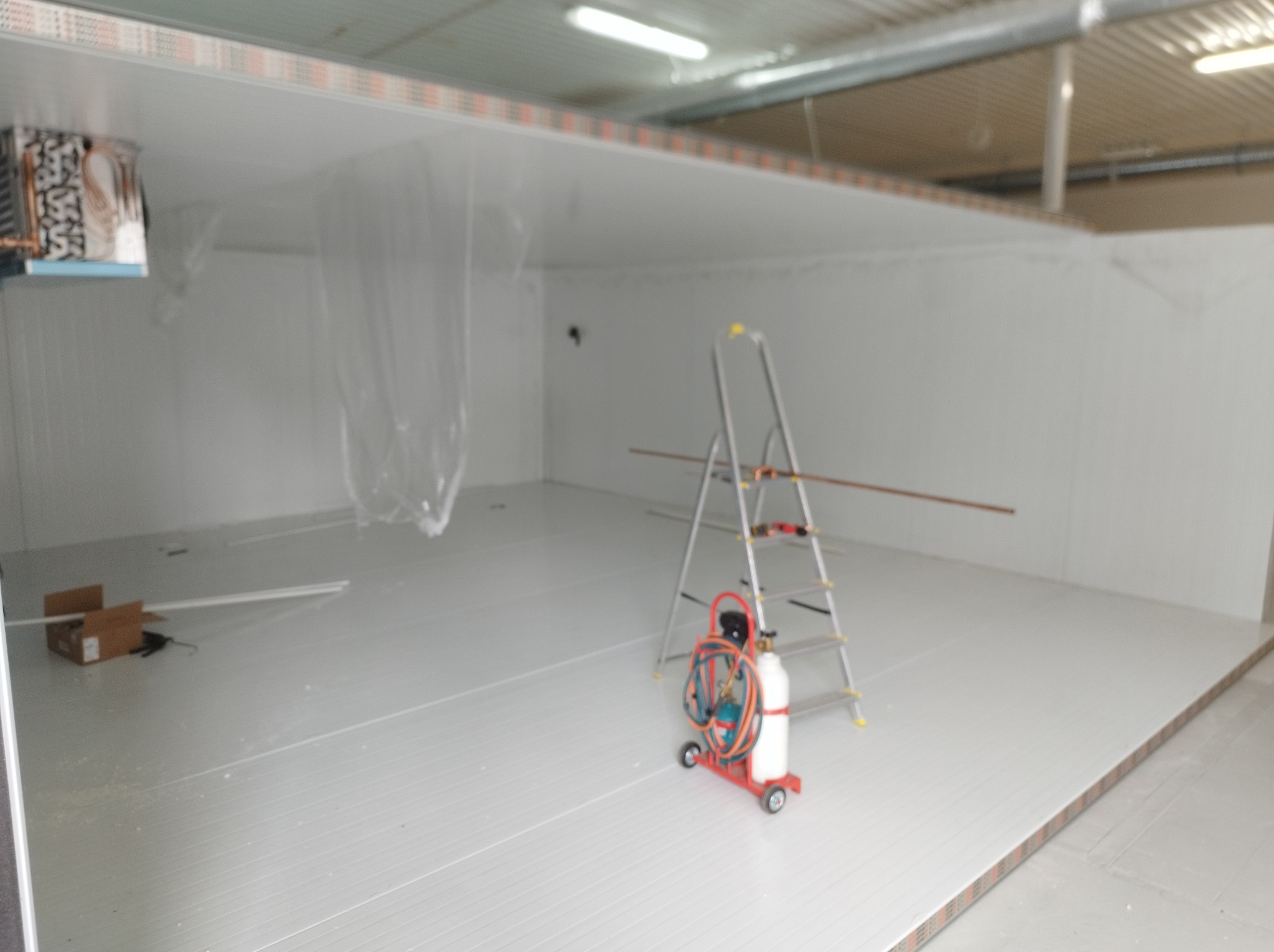 Wnętrze chłodni w trakcie montażu. Widoczna drabina, butla z gazem i narzędzia. Białe ściany i podłoga z paneli. Oświetlenie sufitowe.