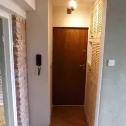 Korytarz po remoncie: odnowiona drewniana podłoga w jodełkę, ściany w odcieniach bieli i szarości, drzwi wejściowe z brązowego drewna, zabudowana szafa z żaluzjowymi drzwiczkami.