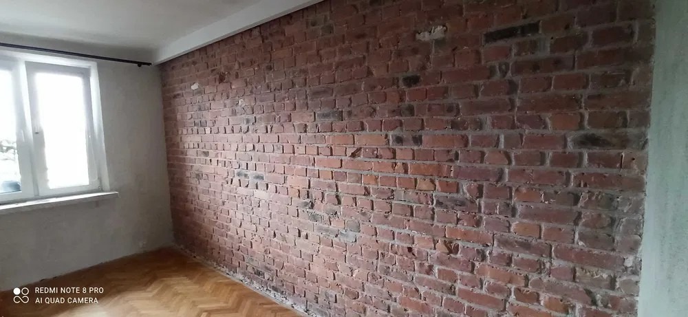 Ceglana ściana w pokoju z parkietem, widoczne nierówności i ubytki w spoinach, okno po lewej stronie z białymi ramami, surowy wystrój.