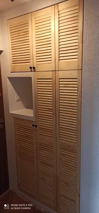 Wysoka, zabudowana szafa z jasnego drewna z lamelkowymi drzwiczkami i czarnymi gałkami, wbudowana we wnękę w przedpokoju z półką na drobiazgi.