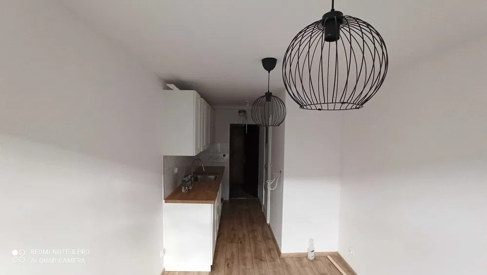 Wnętrze po malowaniu: Białe ściany, drewniana podłoga, minimalistyczna biała zabudowa kuchenna z drewnianym blatem, dwie czarne lampy druciane.