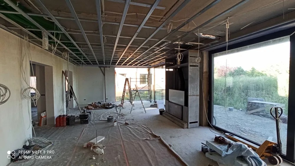 Wnętrze w stanie surowym z widoczną konstrukcją sufitu podwieszanego z profili metalowych, przygotowane do dalszych prac wykończeniowych, z widocznymi przewodami elektrycznymi, oknem tarasowym...