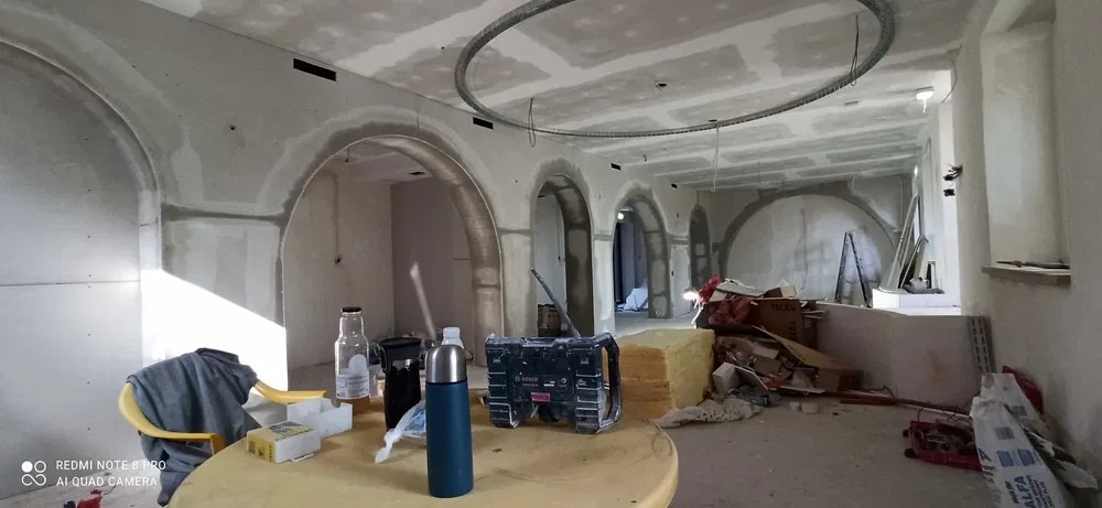 Wnętrze w trakcie wykańczania: ściany z płyt kartonowo-gipsowych z widocznymi łączeniami, przygotowane do dalszych prac. Sufit z elementami dekoracyjnymi. Na pierwszym planie stół z narzędziami...