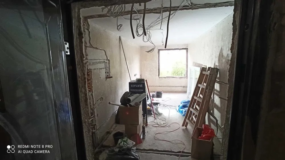 Wnętrze pokoju w trakcie remontu: surowe ściany, odsłonięte przewody elektryczne, drabina oparta o ścianę, kartony i materiały budowlane w nieładzie, widok zza otwartych drzwi.
