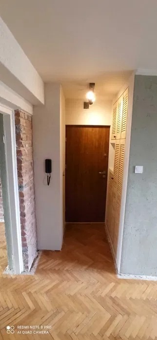 Korytarz po remoncie: odnowiona drewniana podłoga w jodełkę, ściany w odcieniach bieli i szarości, drzwi wejściowe z brązowego drewna, zabudowana szafa z żaluzjowymi drzwiczkami.