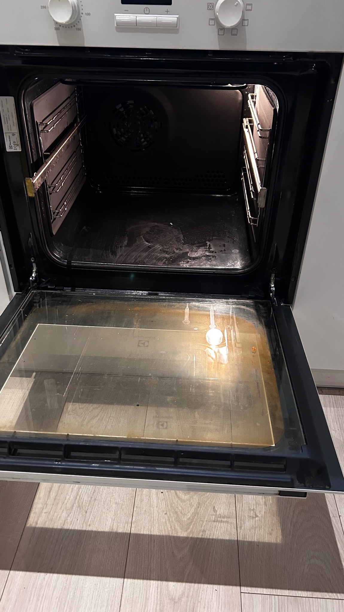 Wnętrze brudnego piekarnika elektrycznego z otwartymi drzwiczkami, widoczne przypalone resztki jedzenia na dnie i szybie, wnętrze wymaga dokładnego wyczyszczenia.