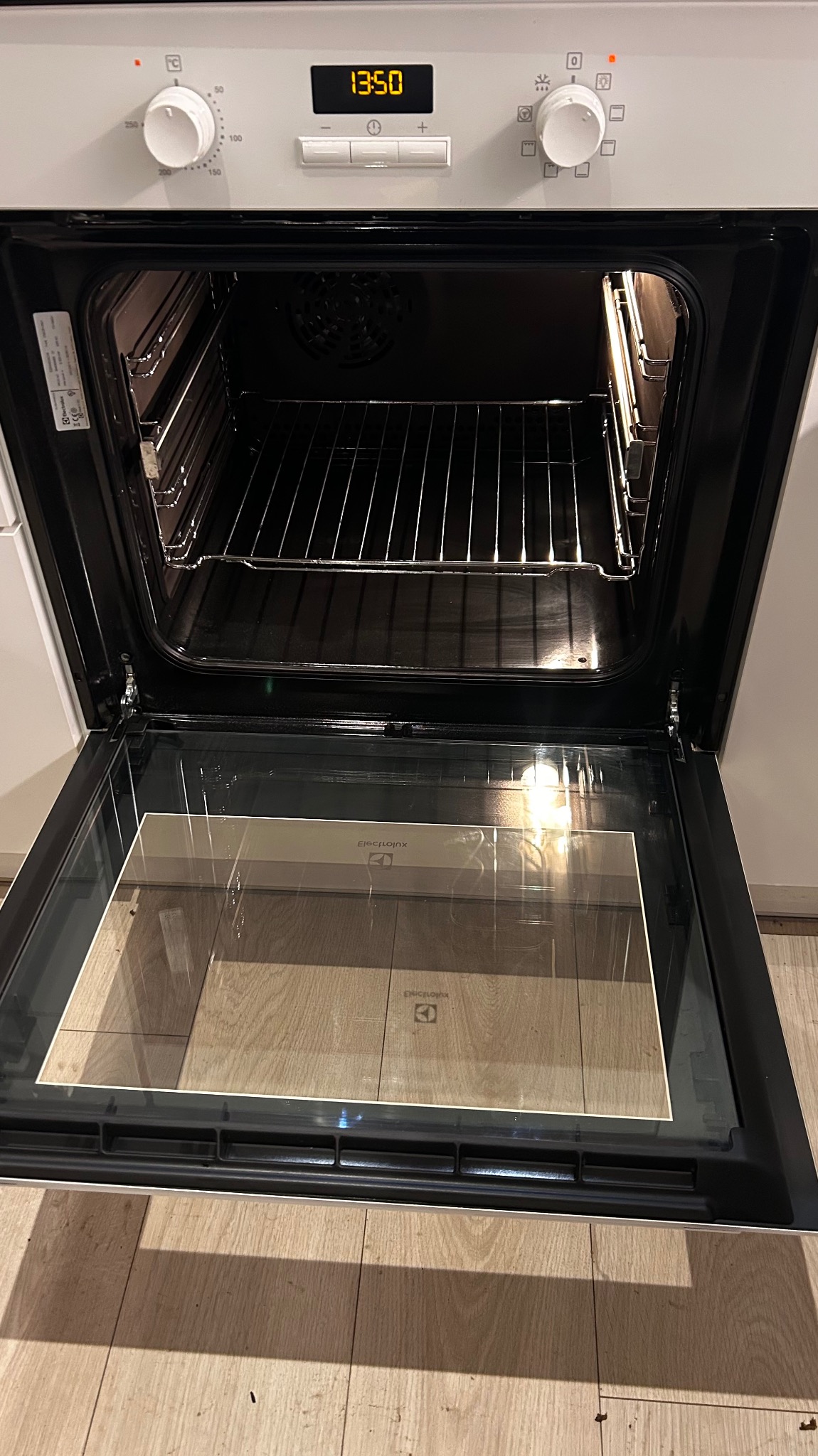 Otwarty piekarnik elektryczny marki Electrolux, widoczne wnętrze z metalowymi prowadnicami, cyfrowy wyświetlacz z godziną 13:50, wnętrze czyste, oświetlone, na podłodze widoczne panele.