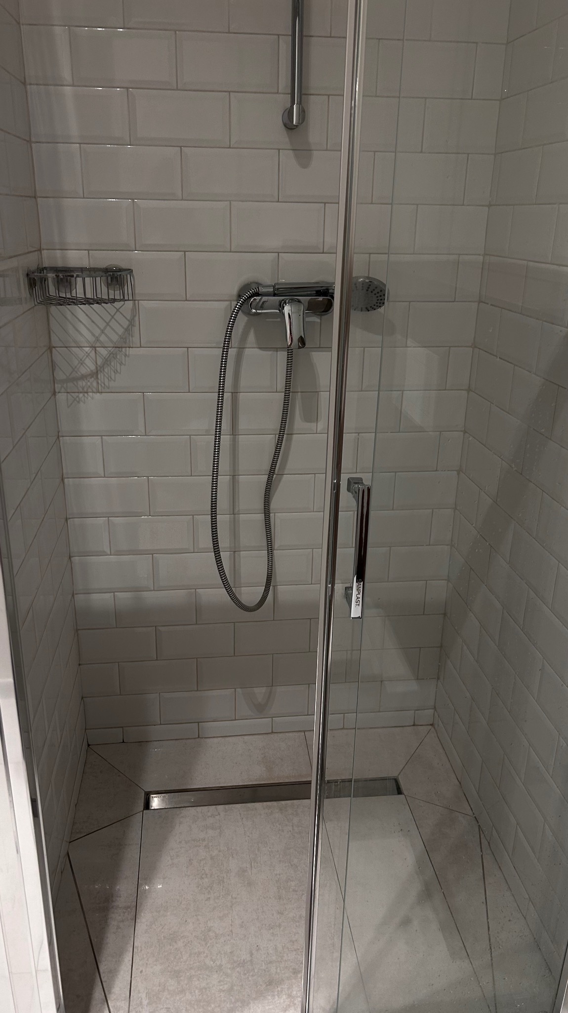 Szczegółowe ujęcie kabiny prysznicowej z chromowaną armaturą, szklanymi drzwiami i odpływem liniowym, wykończonej białymi, prostokątnymi płytkami.