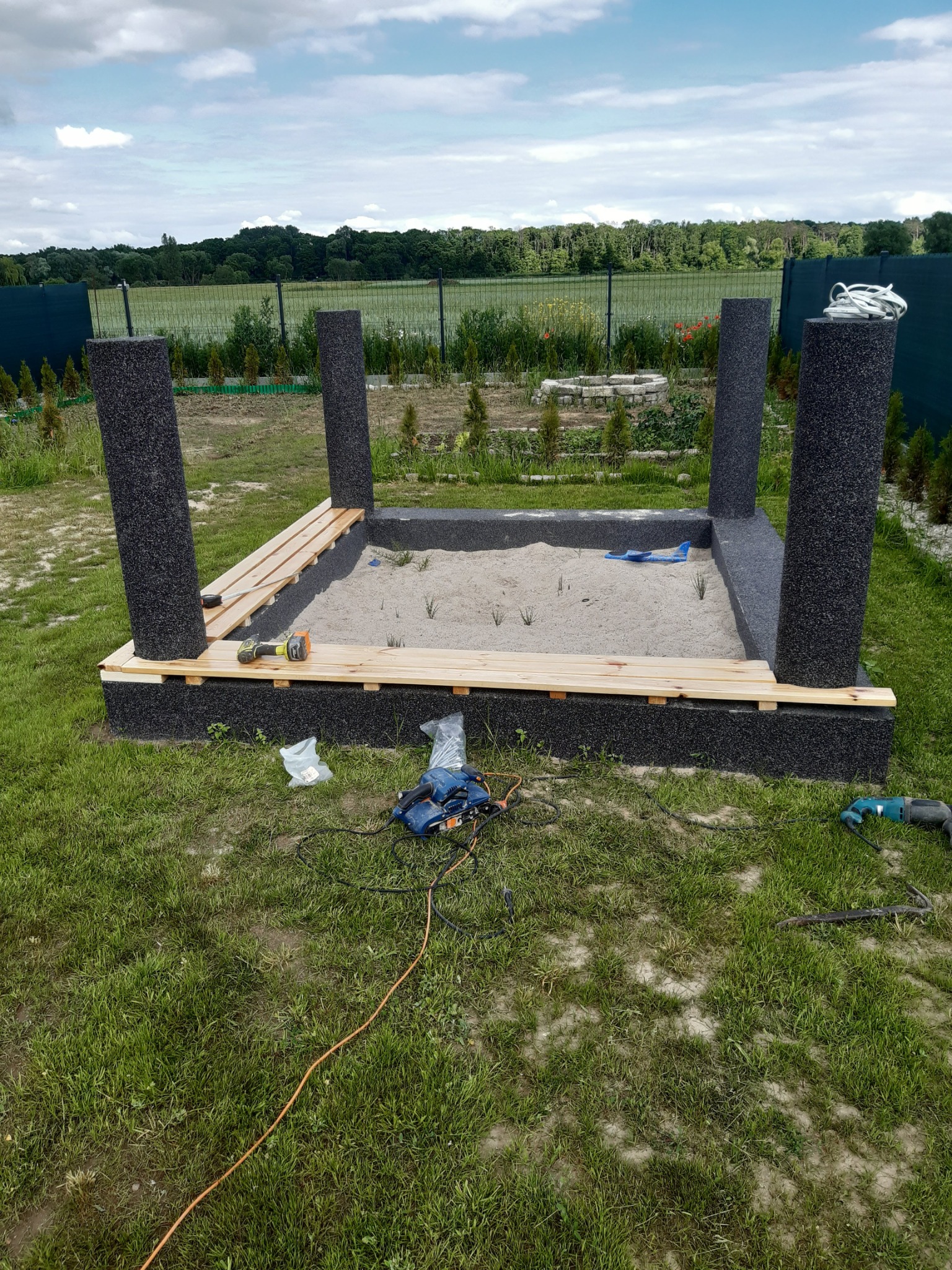 Budowa piaskownicy z czterema czarnymi słupkami i drewnianymi deskami, z narzędziami leżącymi obok na trawie.