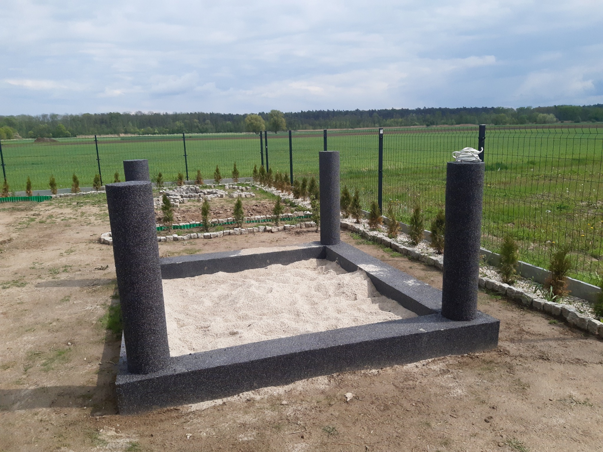 Nowoczesna piaskownica z szarego, granulowanego tworzywa z czterema słupkami w Luboniu, wypełniona piaskiem, otoczona niskimi tujami i siatką ogrodzeniową na tle zielonego pola.