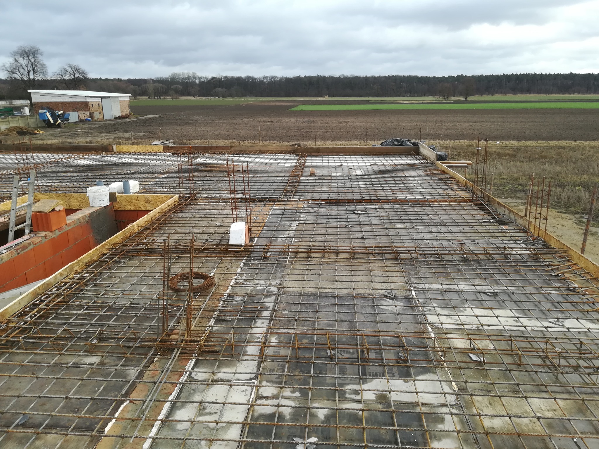 Zbrojenie płyty stropowej na budowie domu jednorodzinnego, widoczne szalunki i wystające pręty zbrojeniowe, w tle krajobraz polny i zabudowania gospodarcze pod pochmurnym niebem.