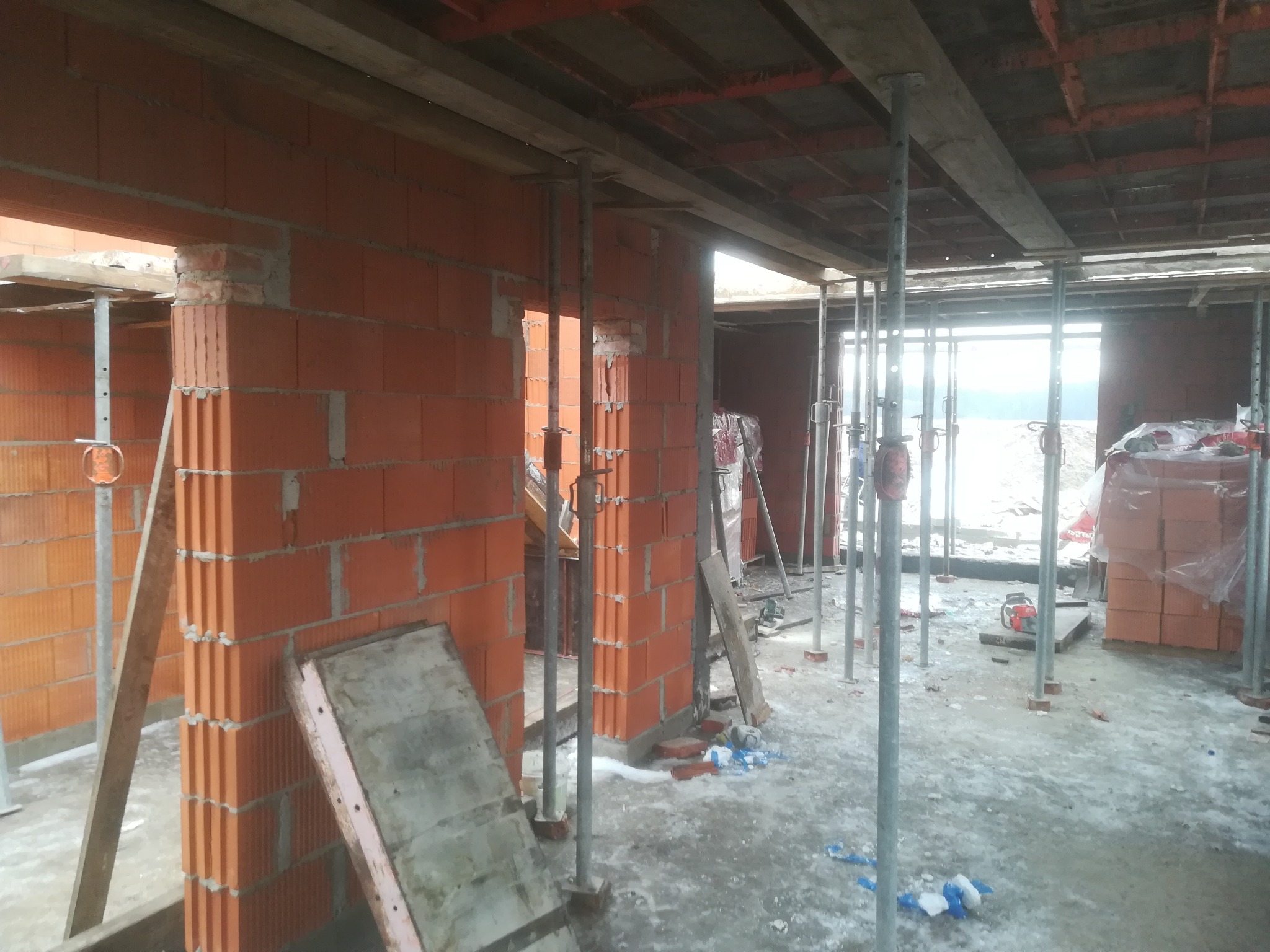 Wnętrze budynku w stanie surowym otwartym, ściany z czerwonej cegły, metalowe podpory stropu, widoczne narzędzia i materiały budowlane.