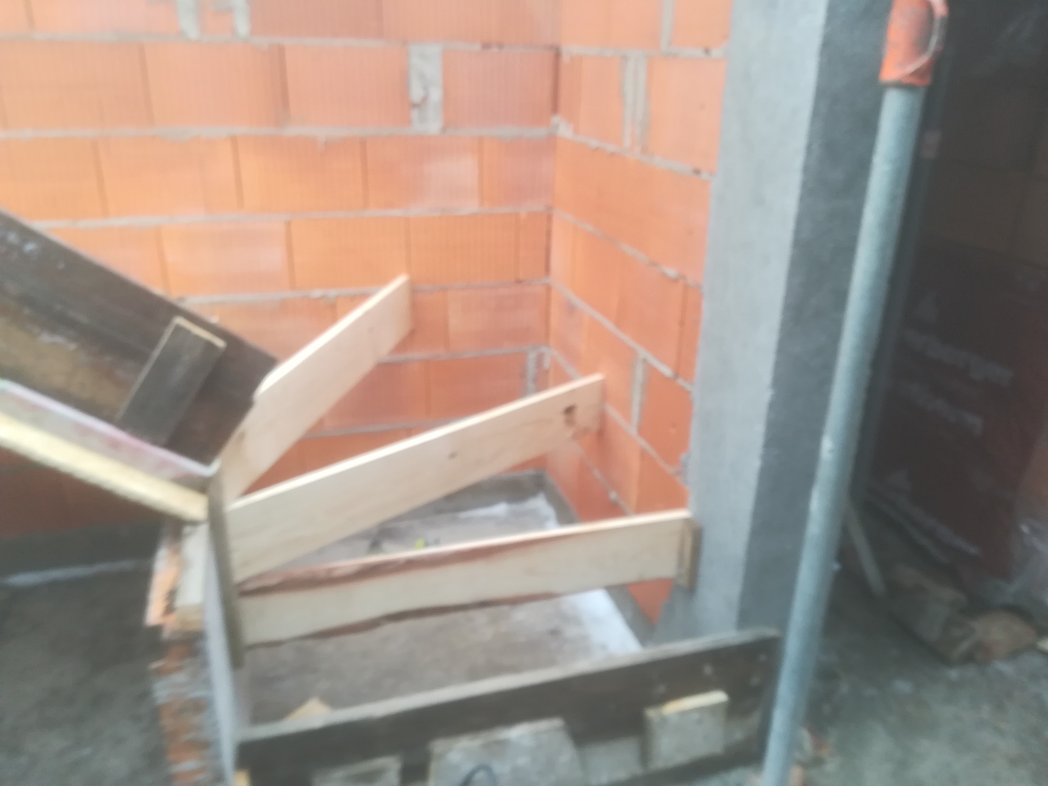 Wewnętrzna konstrukcja schodów z drewnianymi belkami, przymocowana do ściany z czerwonej cegły i betonowego filaru, widoczna w surowym stanie podczas budowy.