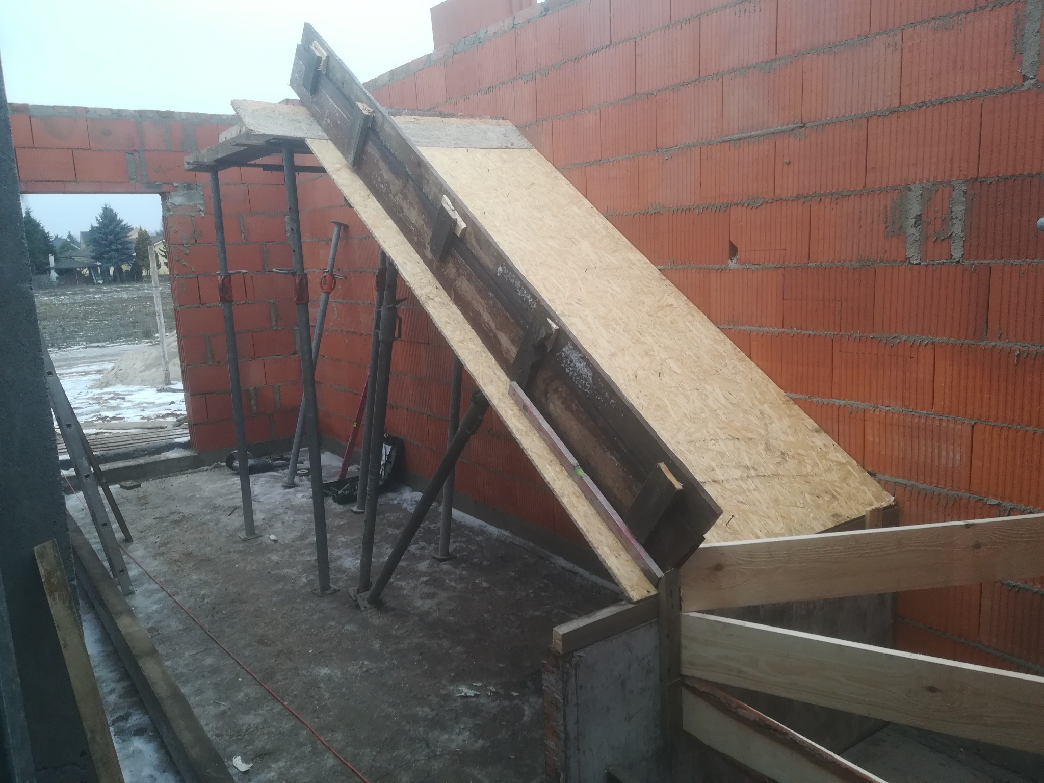 Budowa domu w trakcie: widok na ceglaną ścianę i drewniany szalunek pod kątem, wsparty na stalowych podporach, z widocznym krajobrazem za oknem.