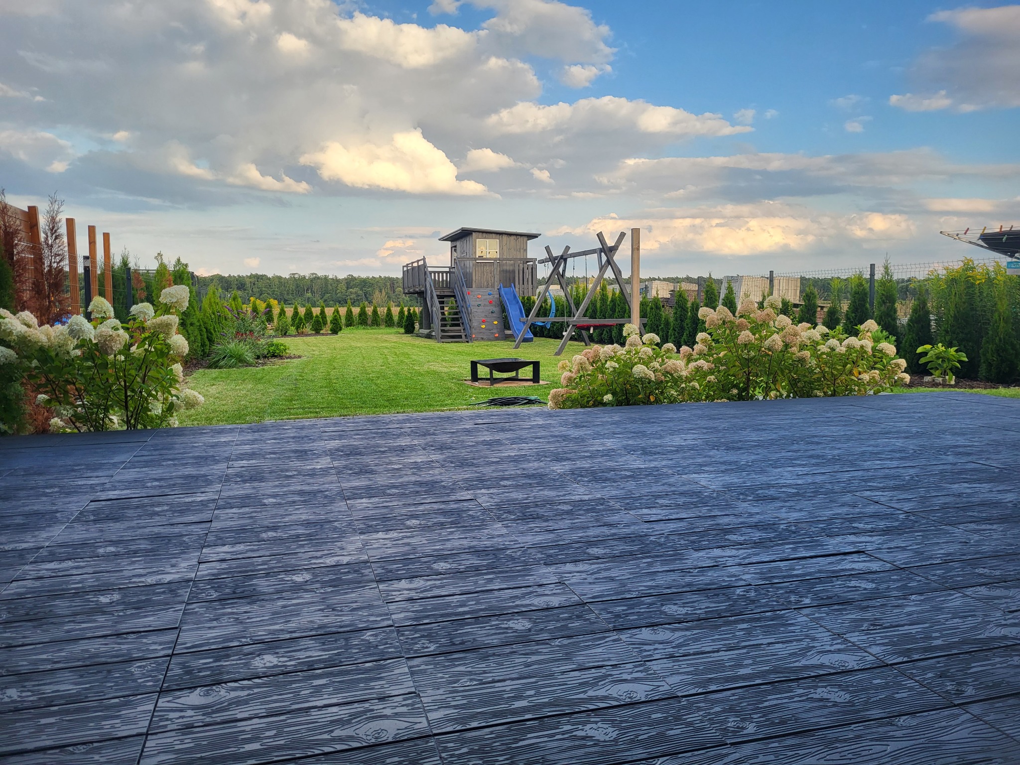 Drewnopodobna, ciemna deska tarasowa prowadząca do zadbanego ogrodu z placem zabaw i bujną roślinnością.