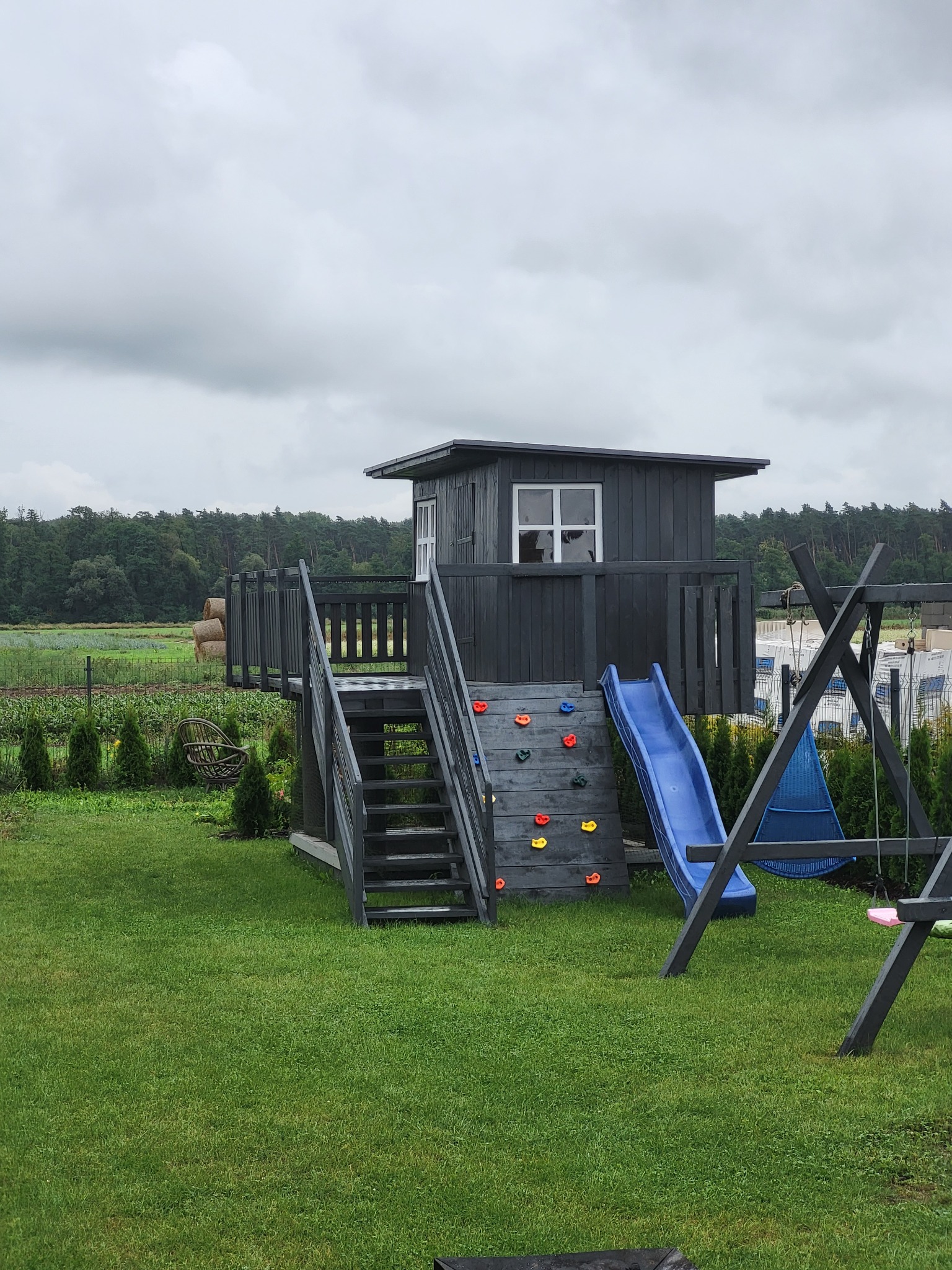 Drewniany plac zabaw z domkiem na platformie, schodami, ścianką wspinaczkową i zjeżdżalnią, umieszczony na zadbanym trawniku na tle pola i lasu.