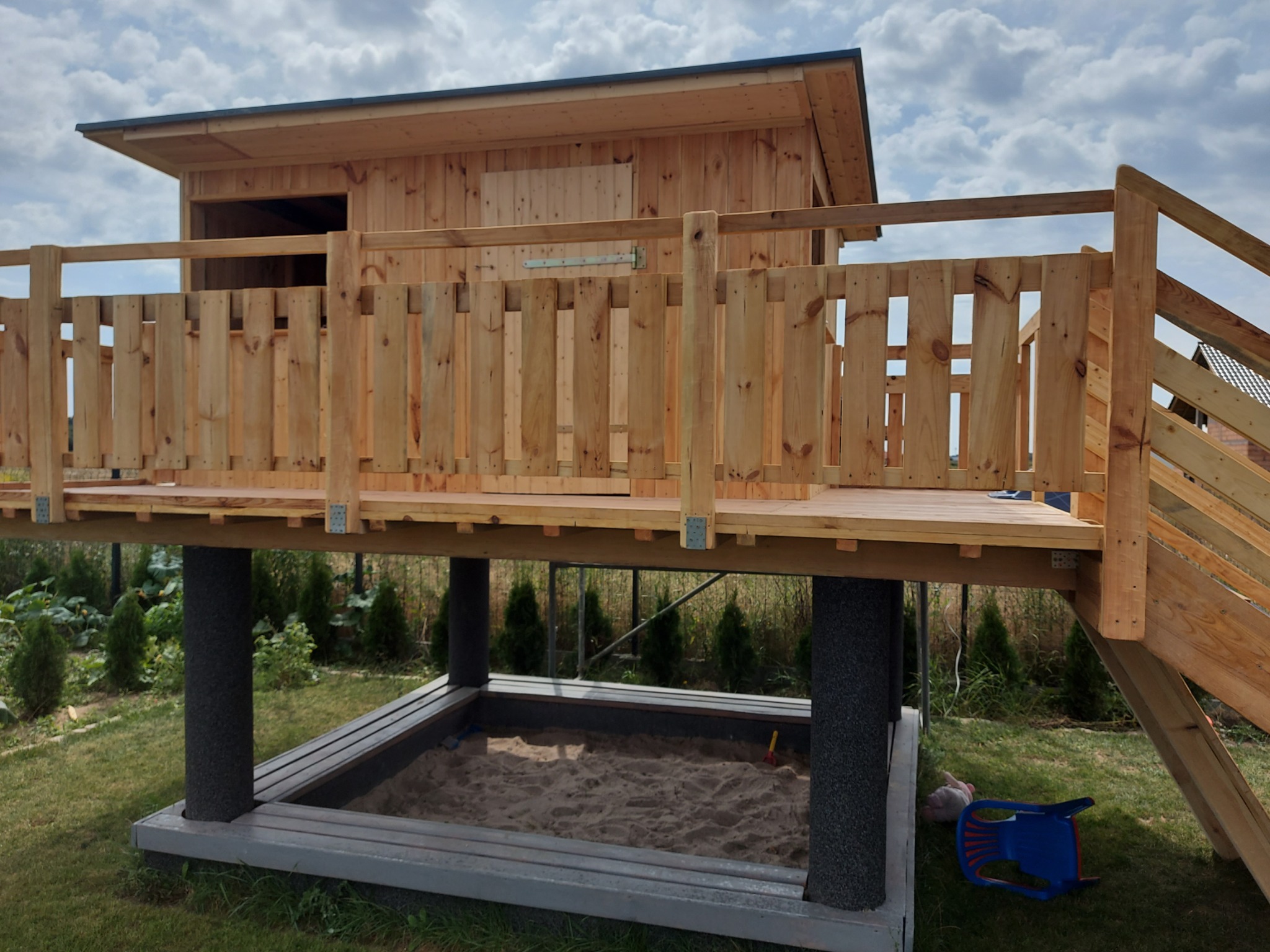 Drewniany domek na palach z piaskownicą poniżej, otoczony zielenią i częściowo zachmurzonym niebem.